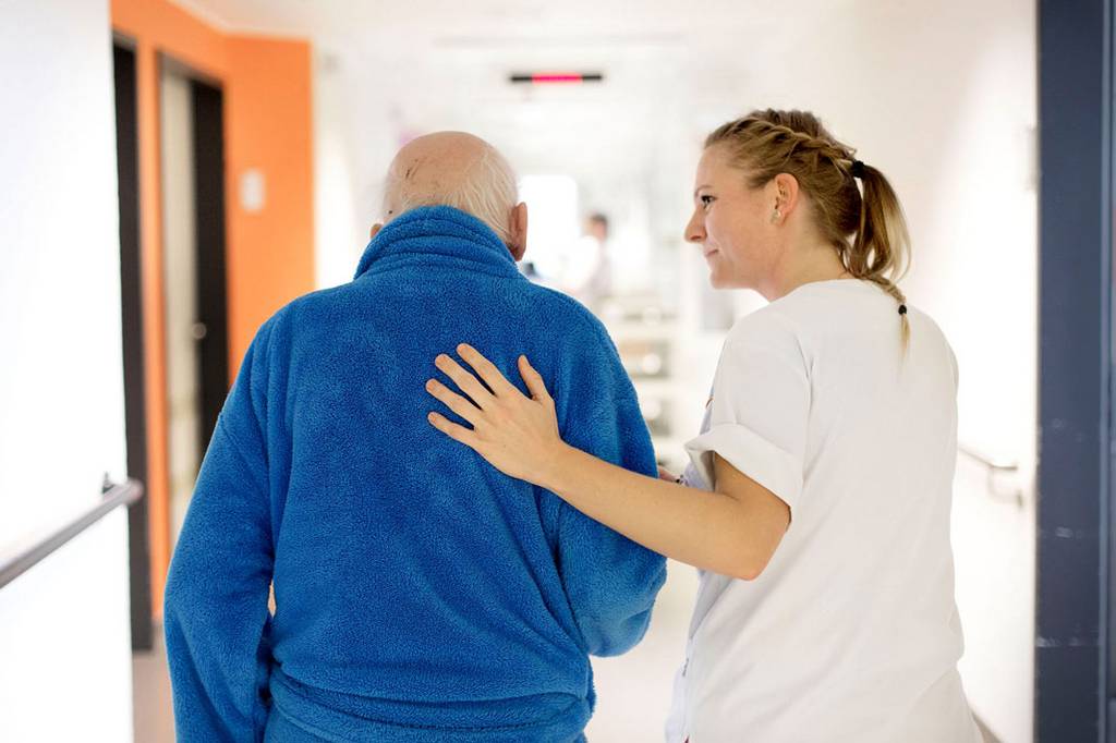 Ein älterer Mann in einem Bademantel wird von einer Krankenpflegerin durch einen Krankenhausflur begleitet. Die Krankenpflegerin hat hierbei eine Hand auf seinem Rücken.
